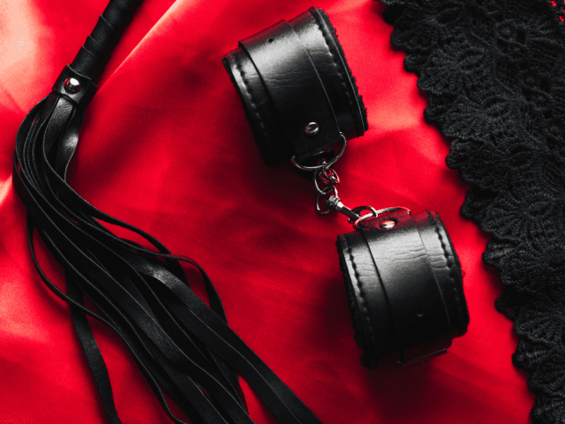 Algemas de couro conectadas por corrente e um flogger sobre tecido vermelho e renda preta, acessórios usados em práticas BDSM.
